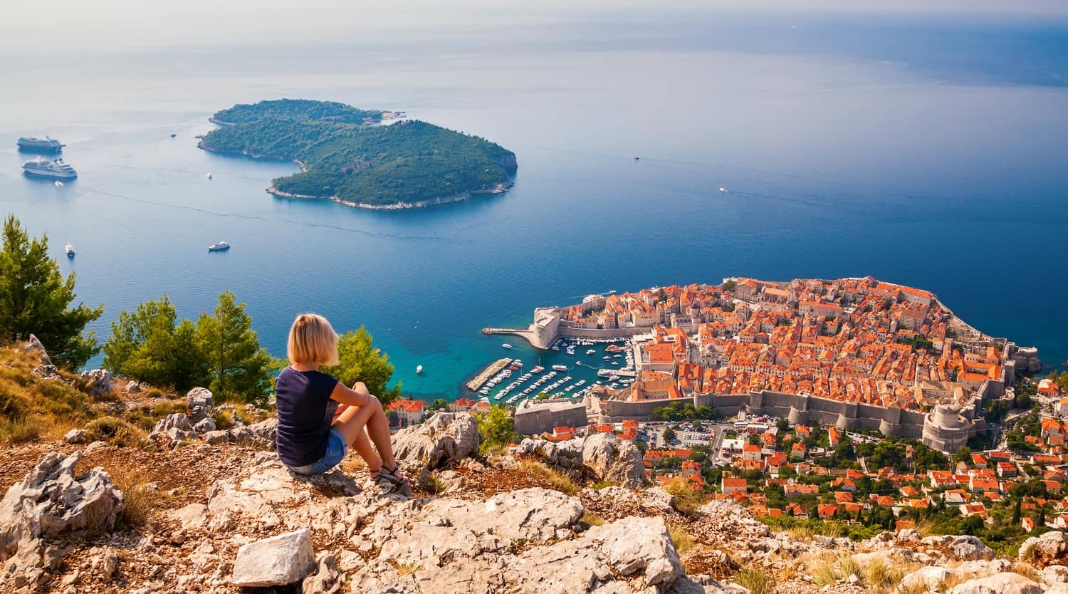 Girl overlooking Dubrovnik's Old Town from the top of a cliff. Ocean visible beyond the harbour area, with ferries and a small island.