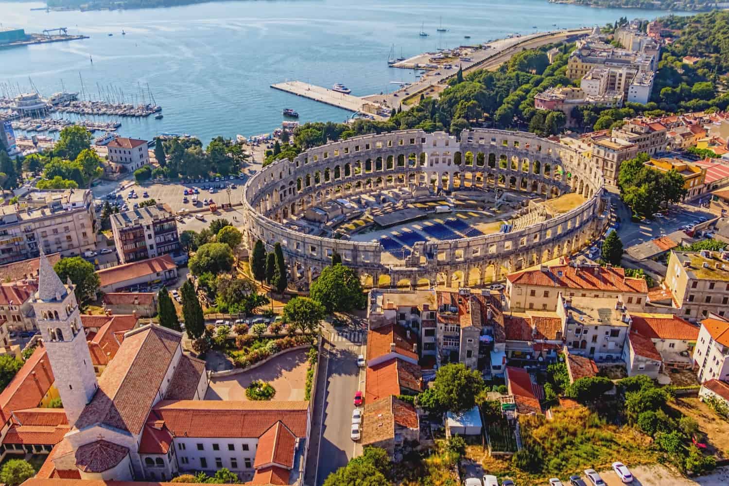 Pula's Roman amphitheatre from above, with red-roofed buildings surrounding it, a church spire visible in front, and a harbour with yachts moored behind.