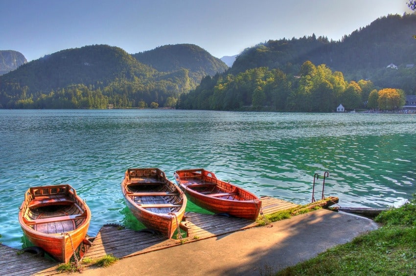 Three small rowing boats tied up at a tiny wooden pier on a lake, with forested hills behind.