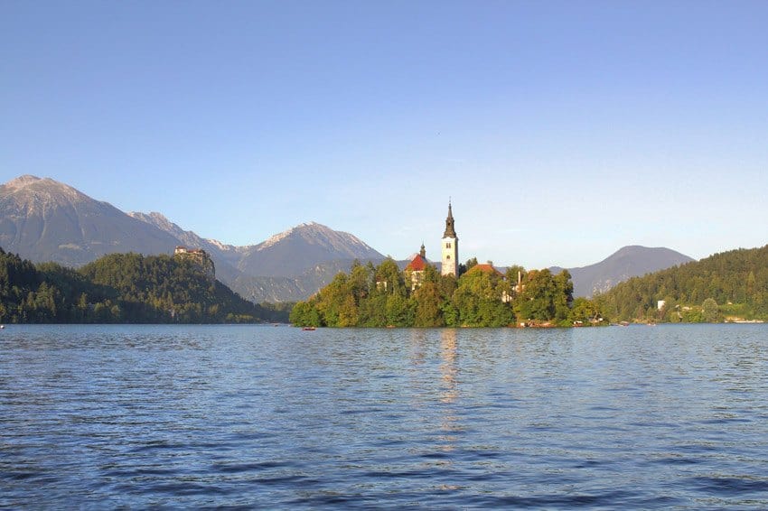 Church on a small island in a lake, with a castle visible on a hill behind and mountains in the distance.