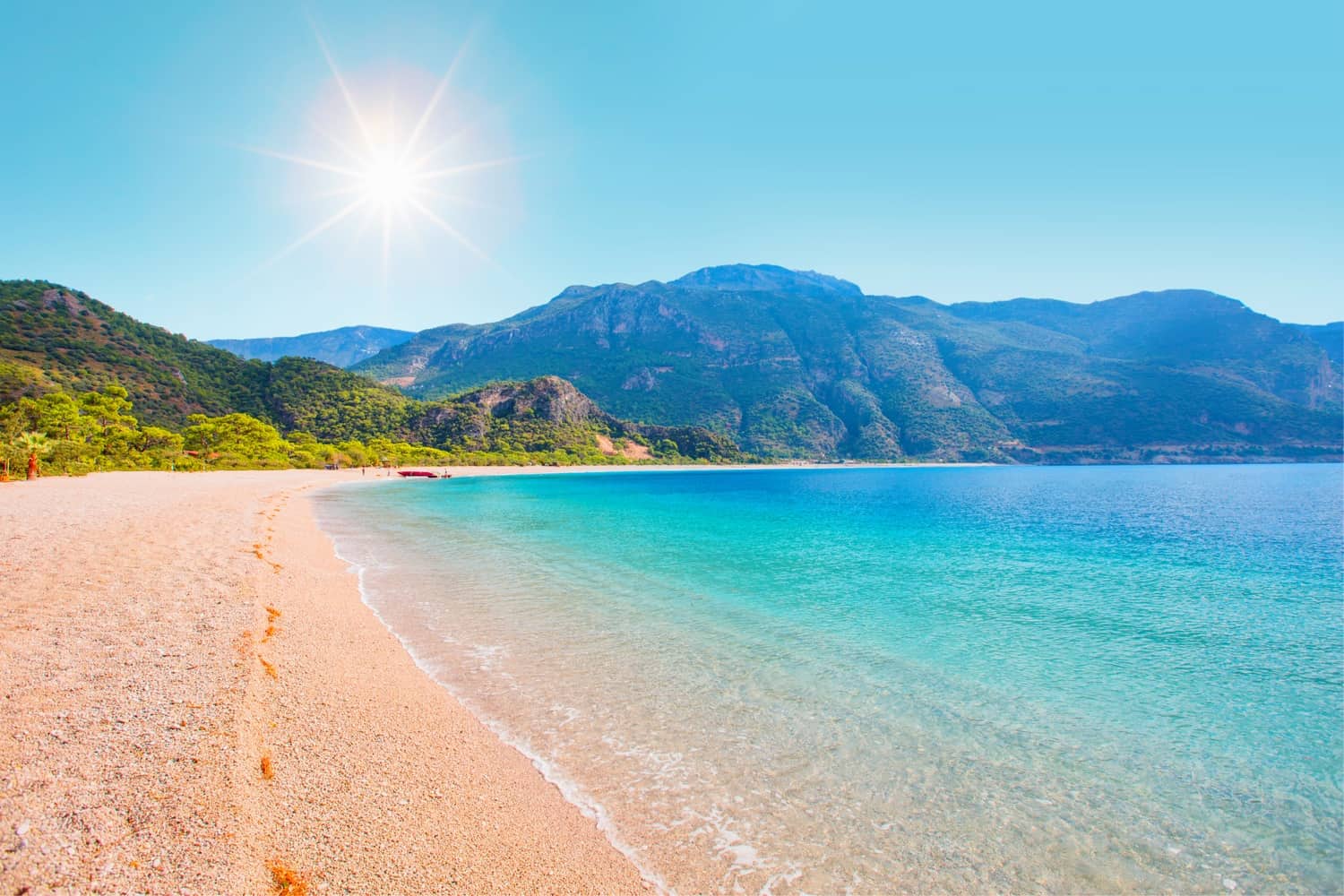 Oludeniz beach in Turkey, with golden sand and small pebbles, surrounded by hills and mountains.