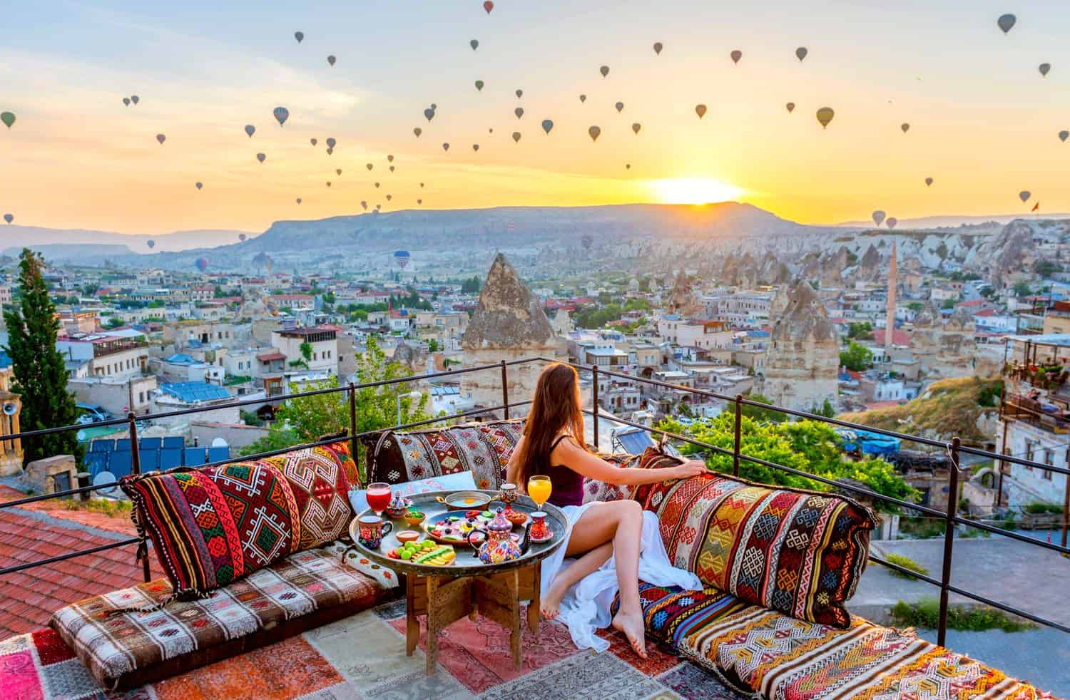 Girl on a rooftop terrace with cushions and breakfast on a tray, turned to watch dozens of hot air balloons rising into the air above the town of Cappadocia