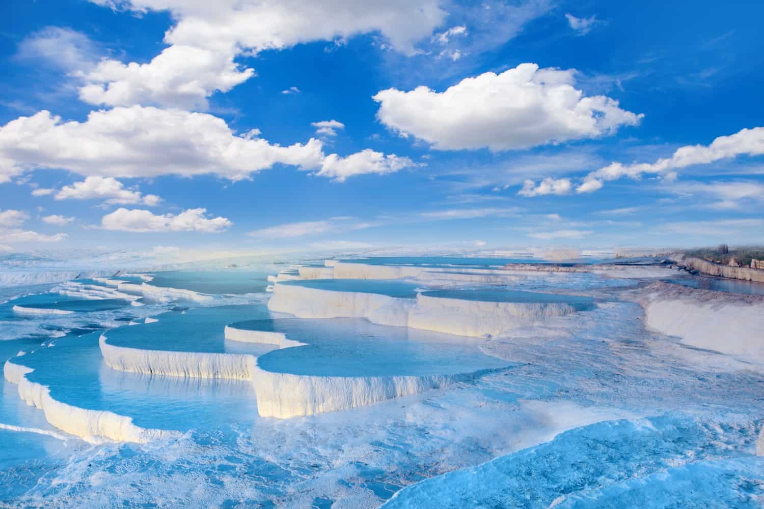 Pamukkale travertines (stepped layers of white limestone rock), each covered in a shallow layer of water with blue sky and white clouds overhead.