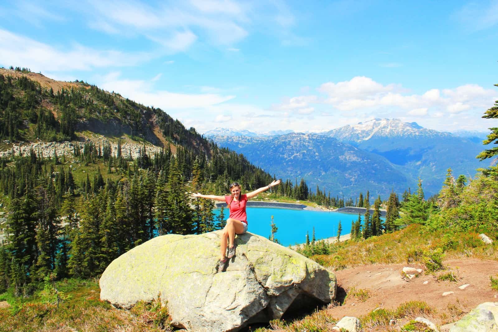 Young woman sitting on a large rock on a grassy hillside, with a small lake behind and mountains in the distance.