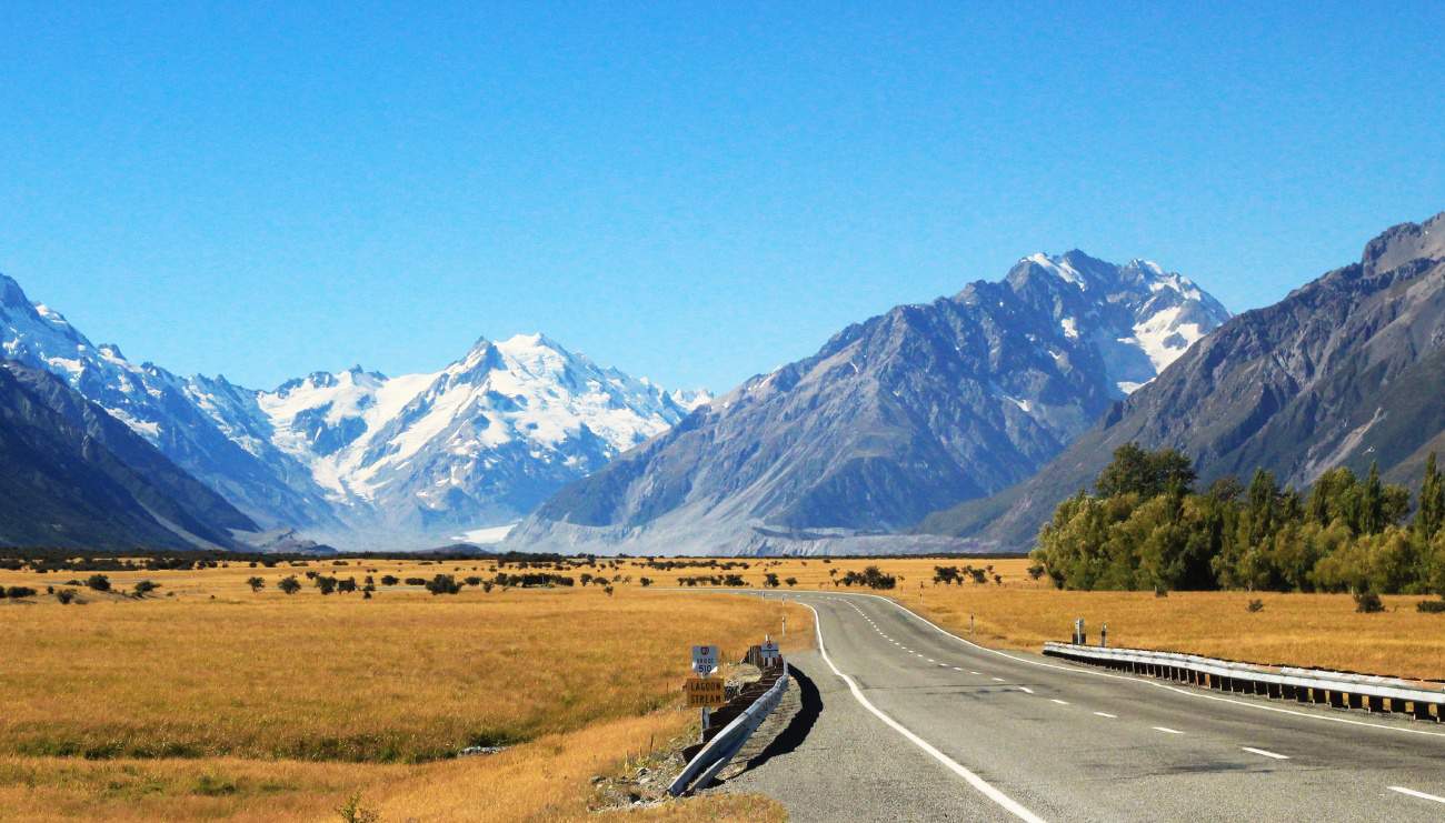 Empty road crossing a small stream in New Zealand, with farmland in the foreground and large mountains in the background.