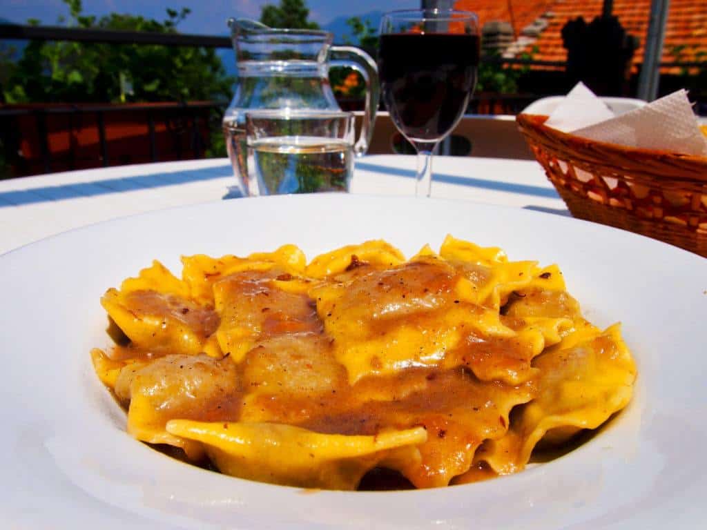 Plate of filled pasta in a white bowl with a glass of red wine and a jug of water behind, on a table on a balcony or terrace.