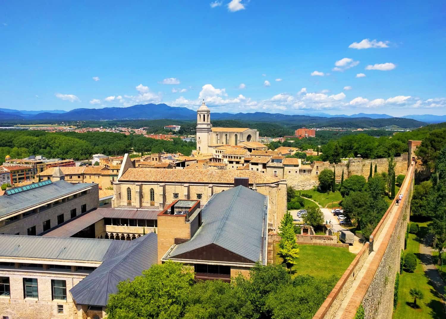 View over and beyond the city walls in Girona, Spain, with buildings inside, many trees outside, and mountains in the distance. A path runs along the top of the walls, with people walking along.