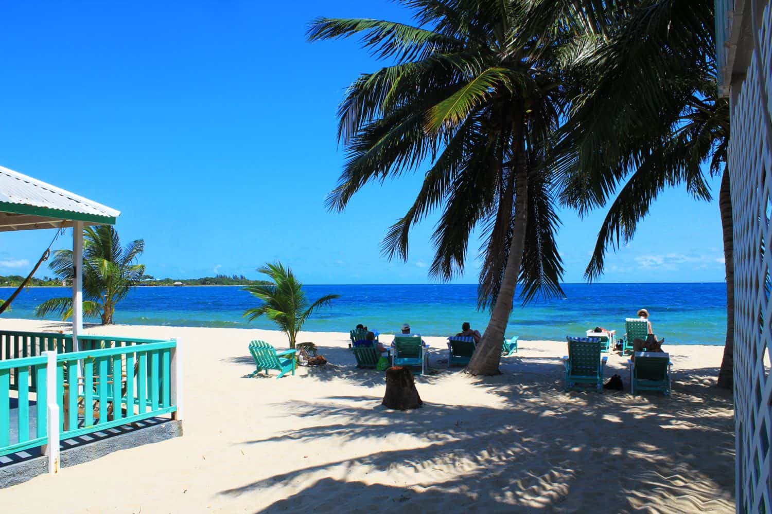 View over a white-sand beach to the ocean, with people sitting on deck chairs under a palm tree, and the side of a terraced eating area to the left.