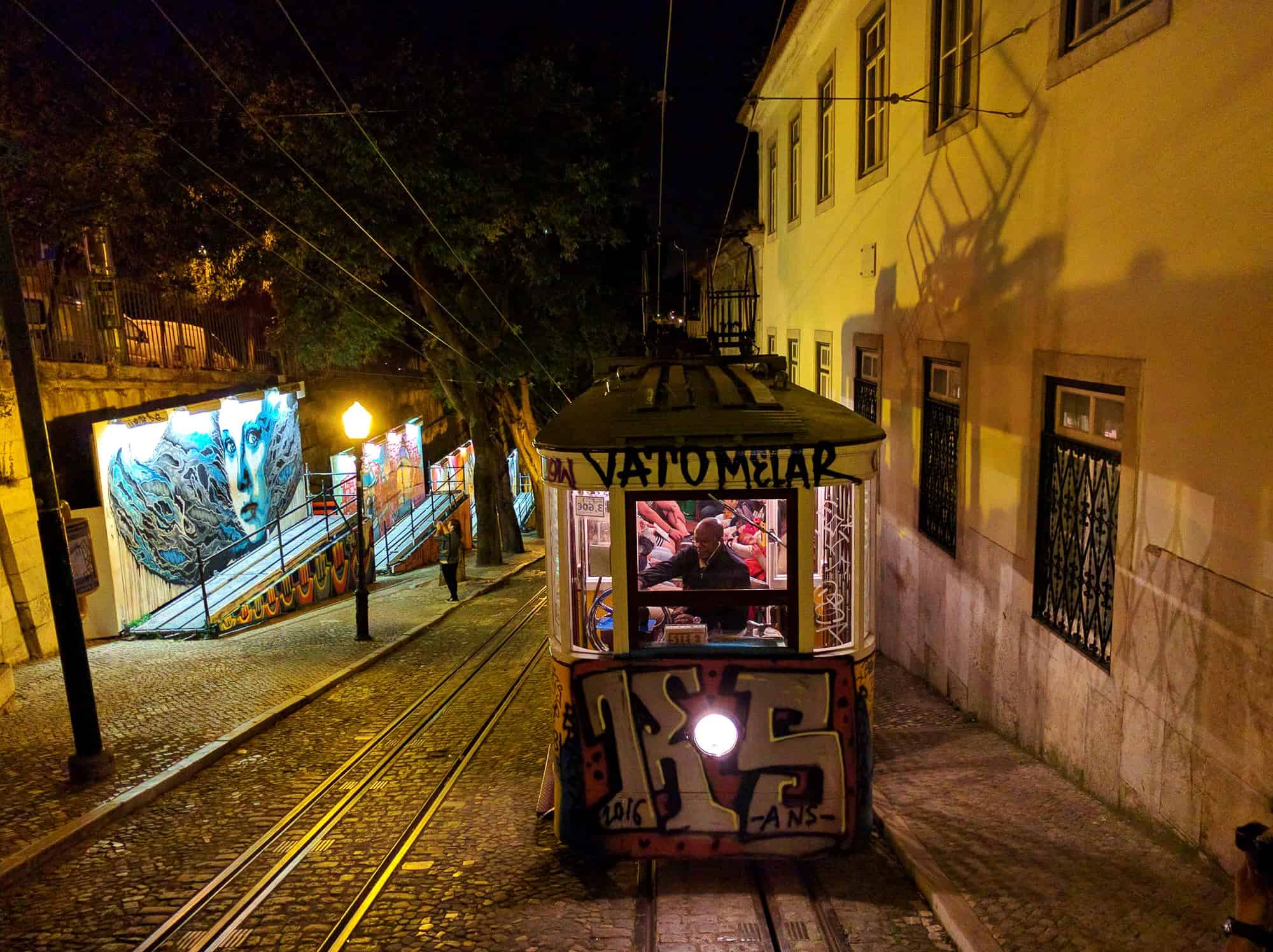 A tram in Lisbon at night, with graffiti on the front and lit up inside as it climbs up a steep cobbled street