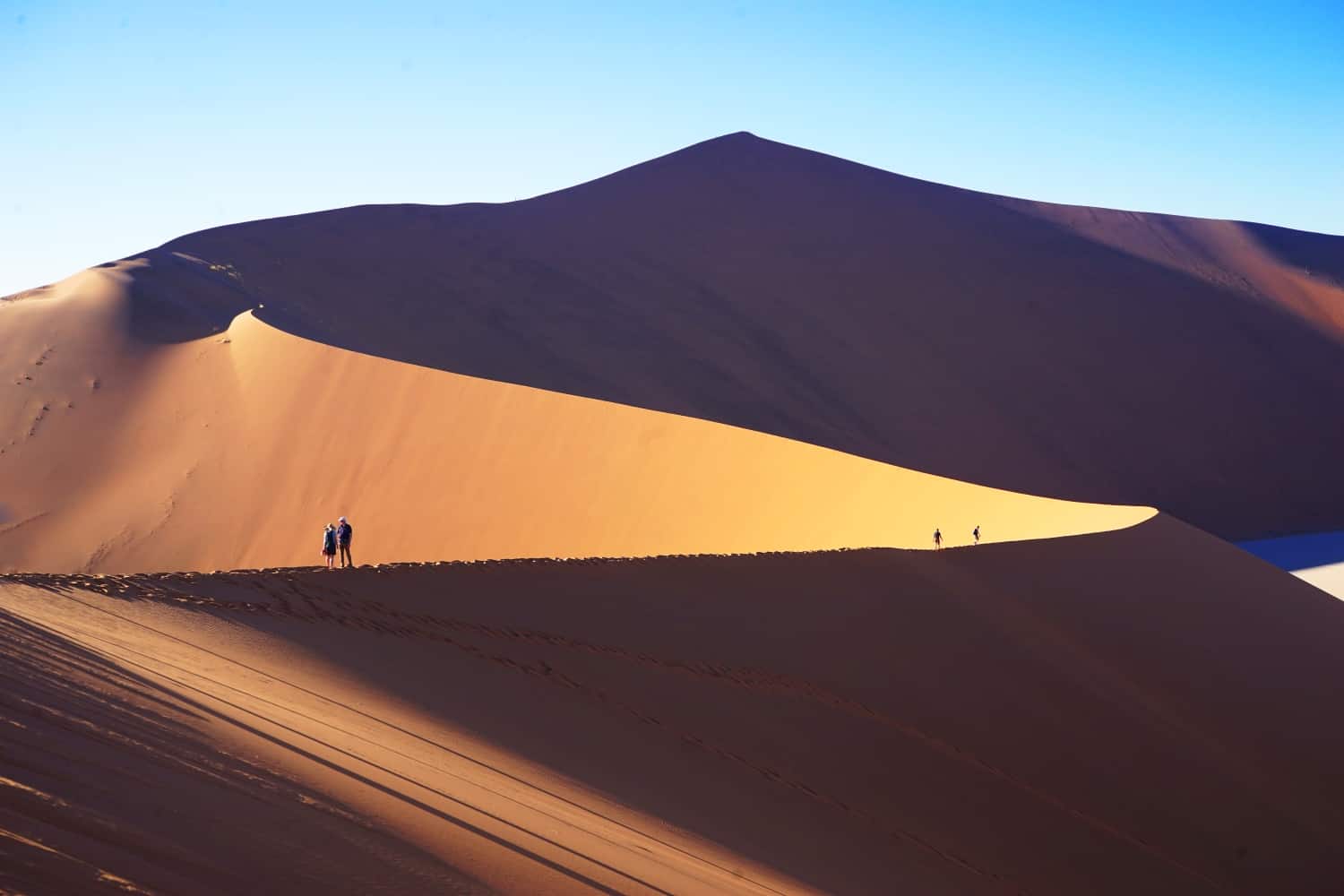 Four people standing and walking along the top of a curved sand dune in Namibia, with another larger dune visible behind.