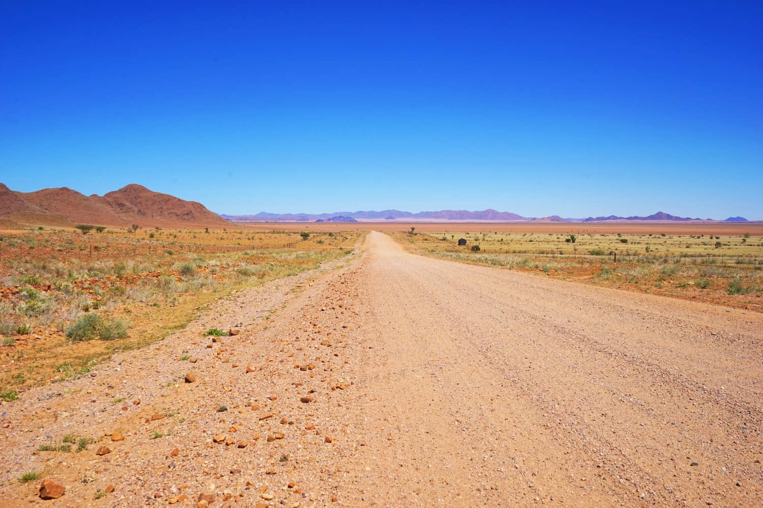 A gravel road in Namibia, with scrubby desert on both sides, hills to the left, and mountains in the distance.