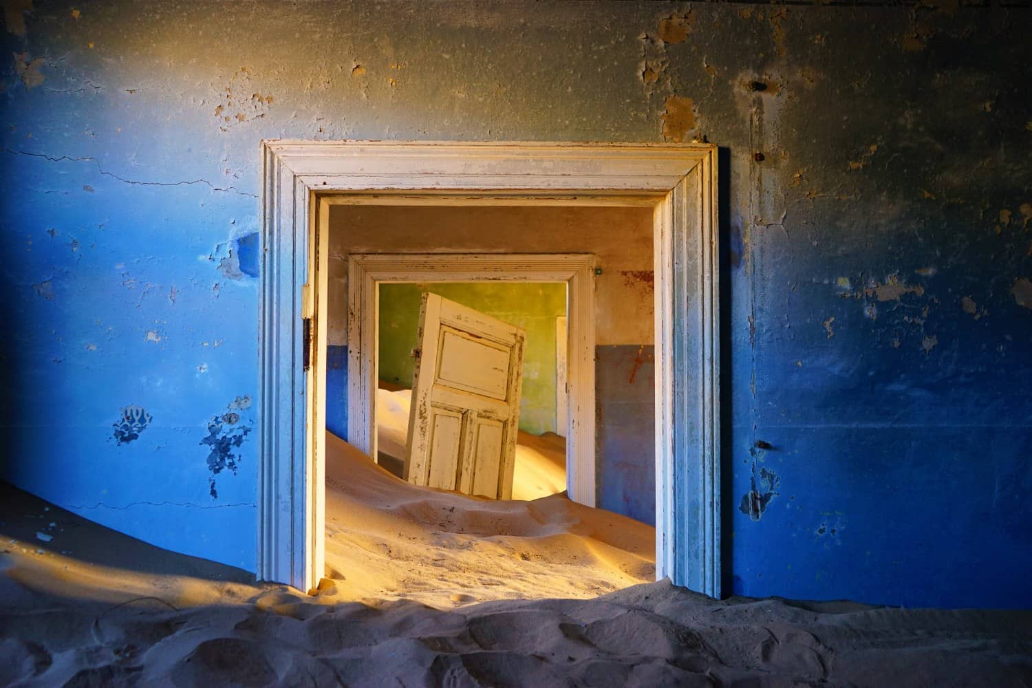 Looking through two doorways with sand covering the floor and climbing up the walls inside a blue-painted house in Kolmanskop, Namibia. A door is standing suspended in the sand, off its hinges.