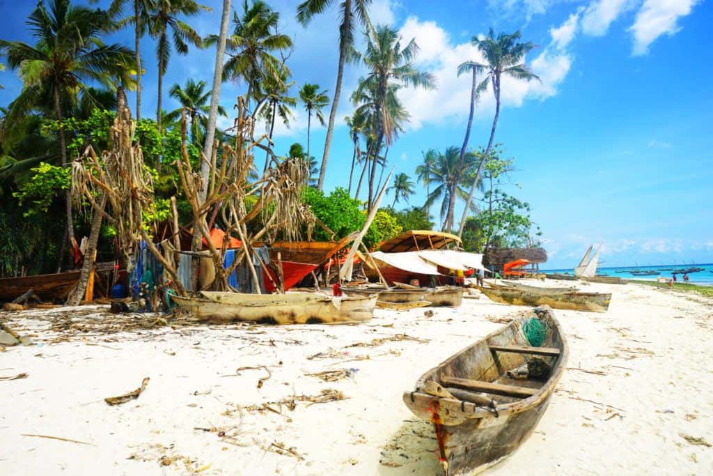 Partially-constructed wooden boats (dhows) on a beach in Nungwi, Zanzibar, with palm trees lining the sand.