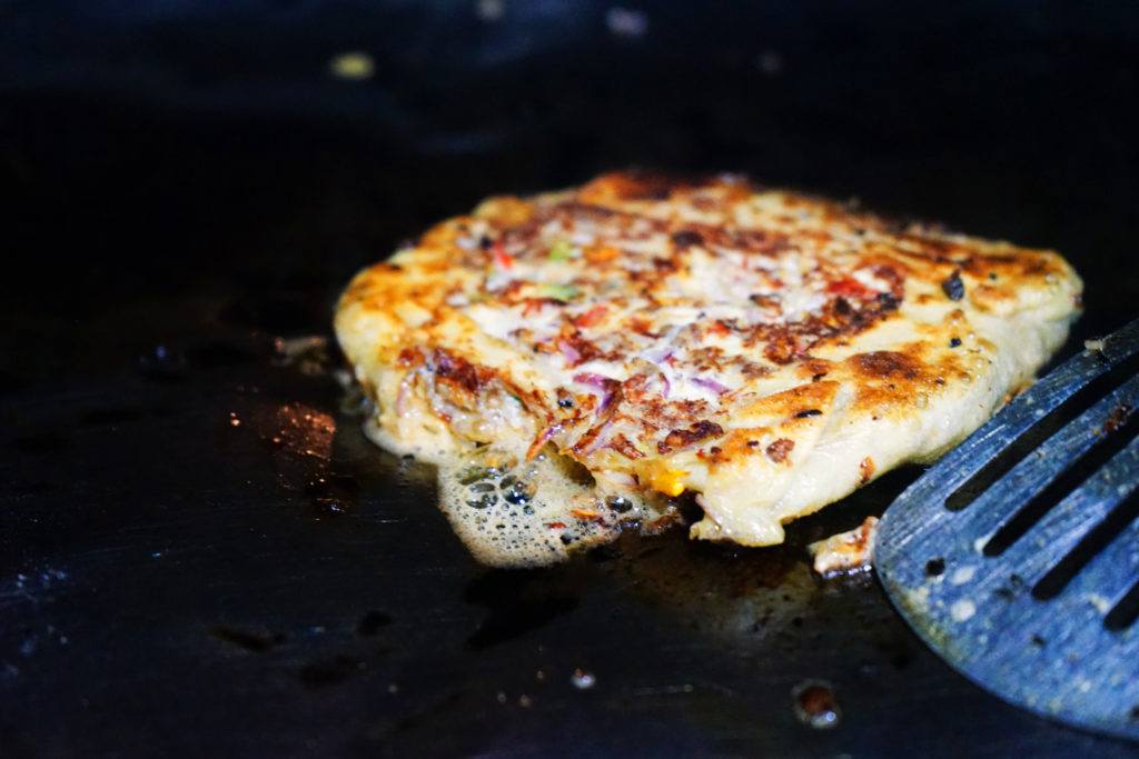 Close-up of a Zanzibar pizza being cooked on a grill, with a partial view of a metal spatula alongside.