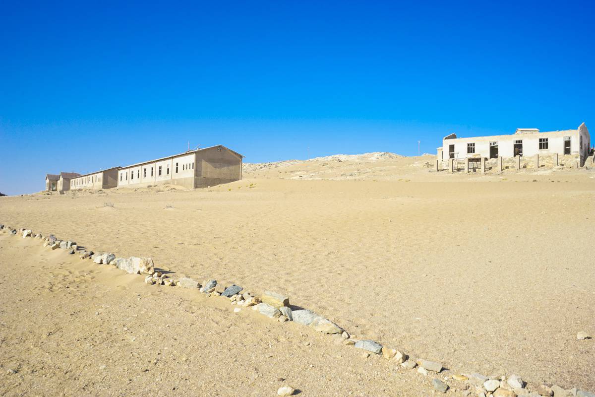 The ghost town complex of Kolmanskop: the pale yellow sand stretches towards the horizon with five abandoned buildings blending into the desert. 