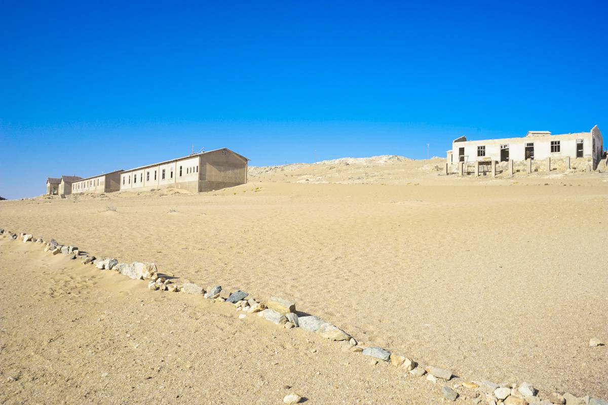 The ghost town complex of Kolmanskop: the pale yellow sand stretches towards the horizon with five abandoned buildings blending into the desert.