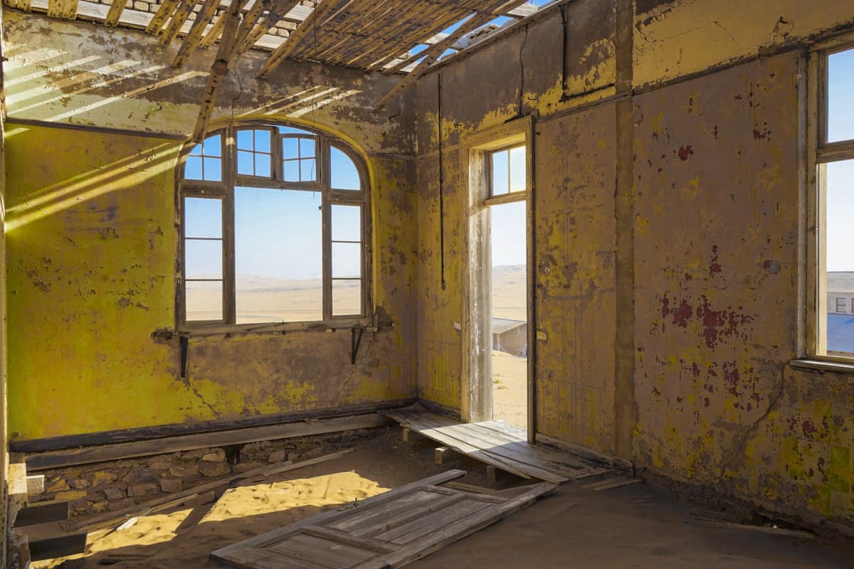 A lime green room with smashed in windows and doors. The floorboards have given way and some of the rafters are falling in, too. Outside the window, you can see endless sand dunes and a bright blue sky