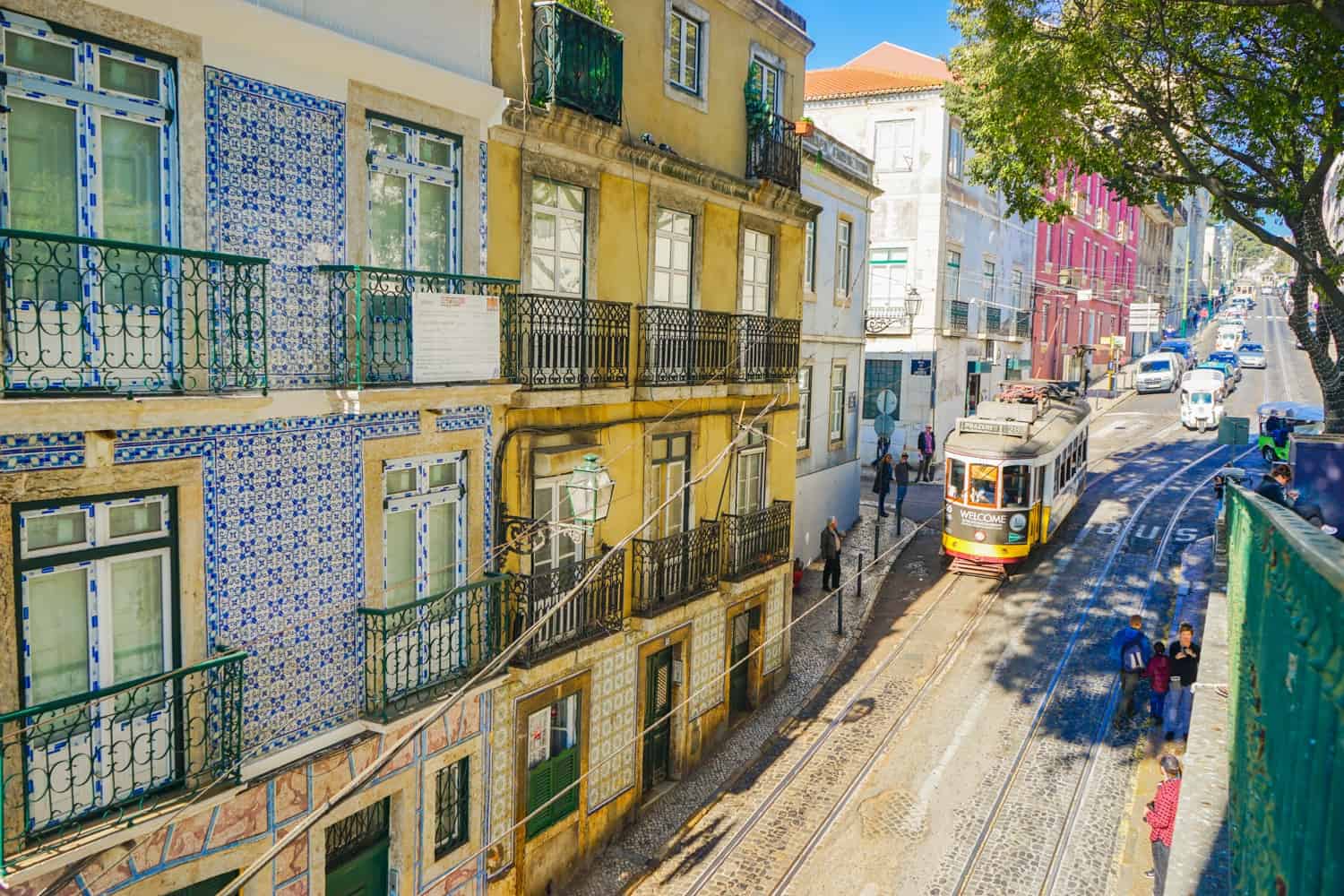 Tram rolling down a typical street in Lisbon, with cobbled streets and pavements, and brightly-coloured tiles on many of the houses