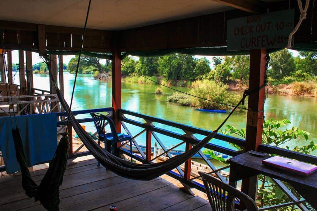 Hammocks strung up on a wooden balcony beside a river, with a thin wooden table and chair on one side. An Apple laptop is sitting closed on the table.