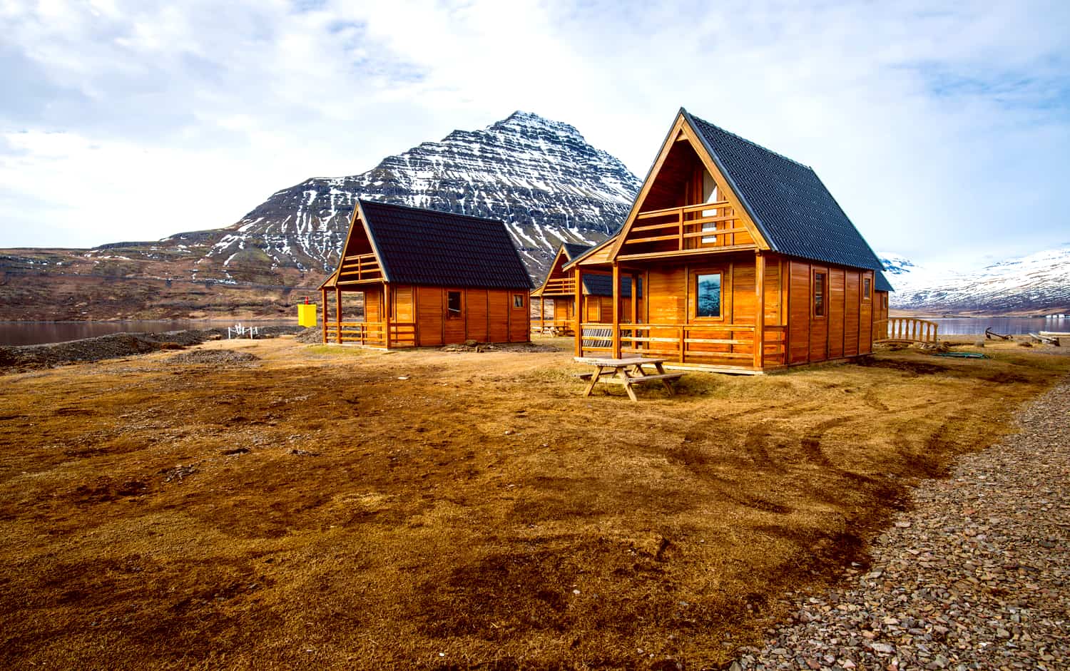 Wooden huts with steeply sloped roofs with a lake and mountains in the background.