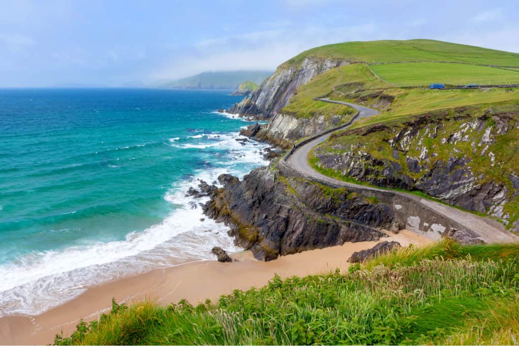 Green hills with steep cliffs above a rocky shoreline and a sandy beach, with a narrow, winding road alongside.