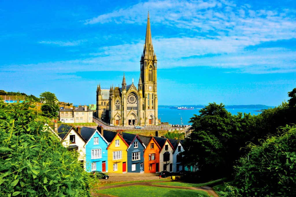 Colorful houses alongside each other on a gentle downhill slope, with a large church on a slight hill behind and ocean visible in the background.