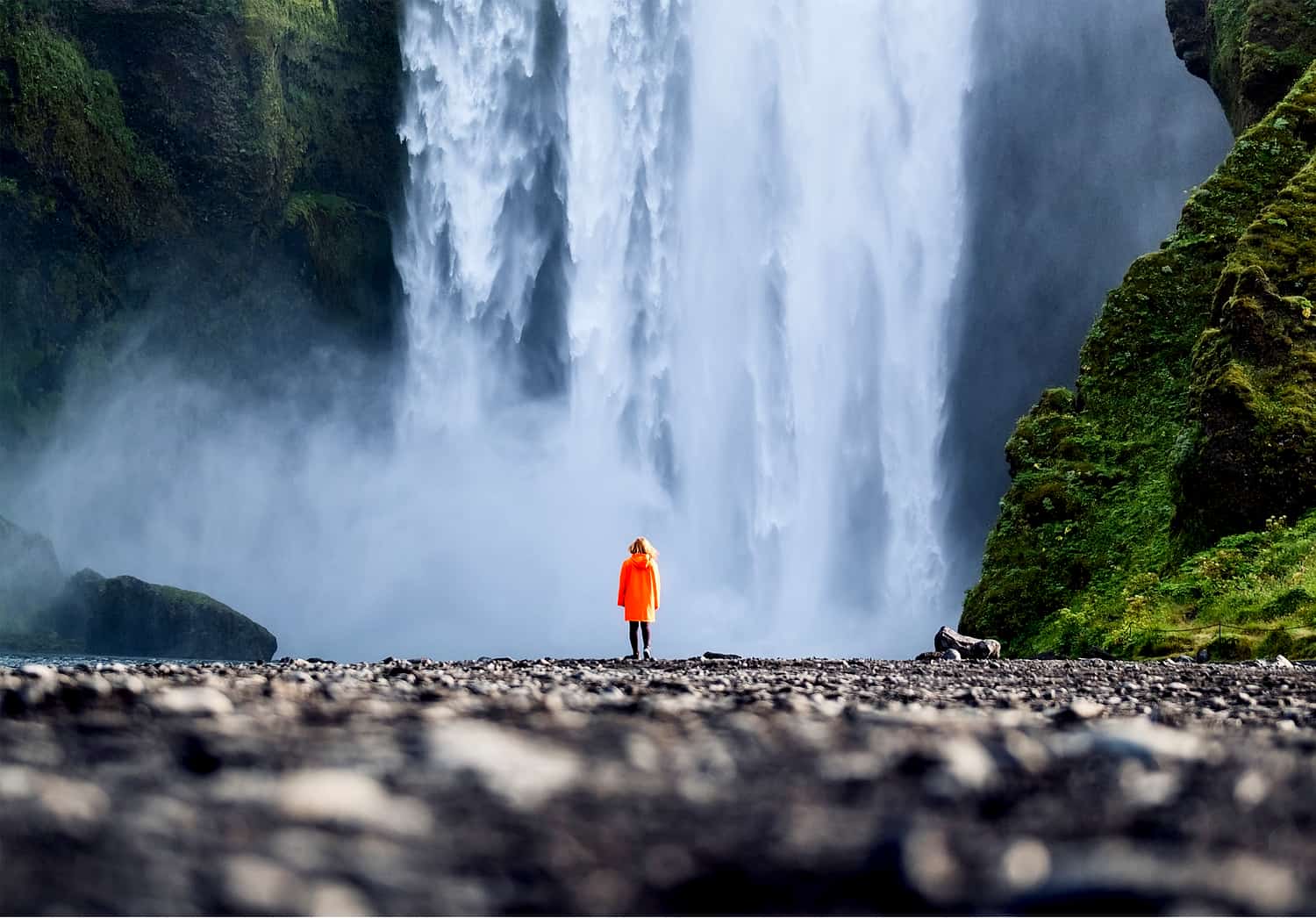 Photo taken at ground level looking towards a woman in an orange jacket standing in front of the base of a large waterfall.