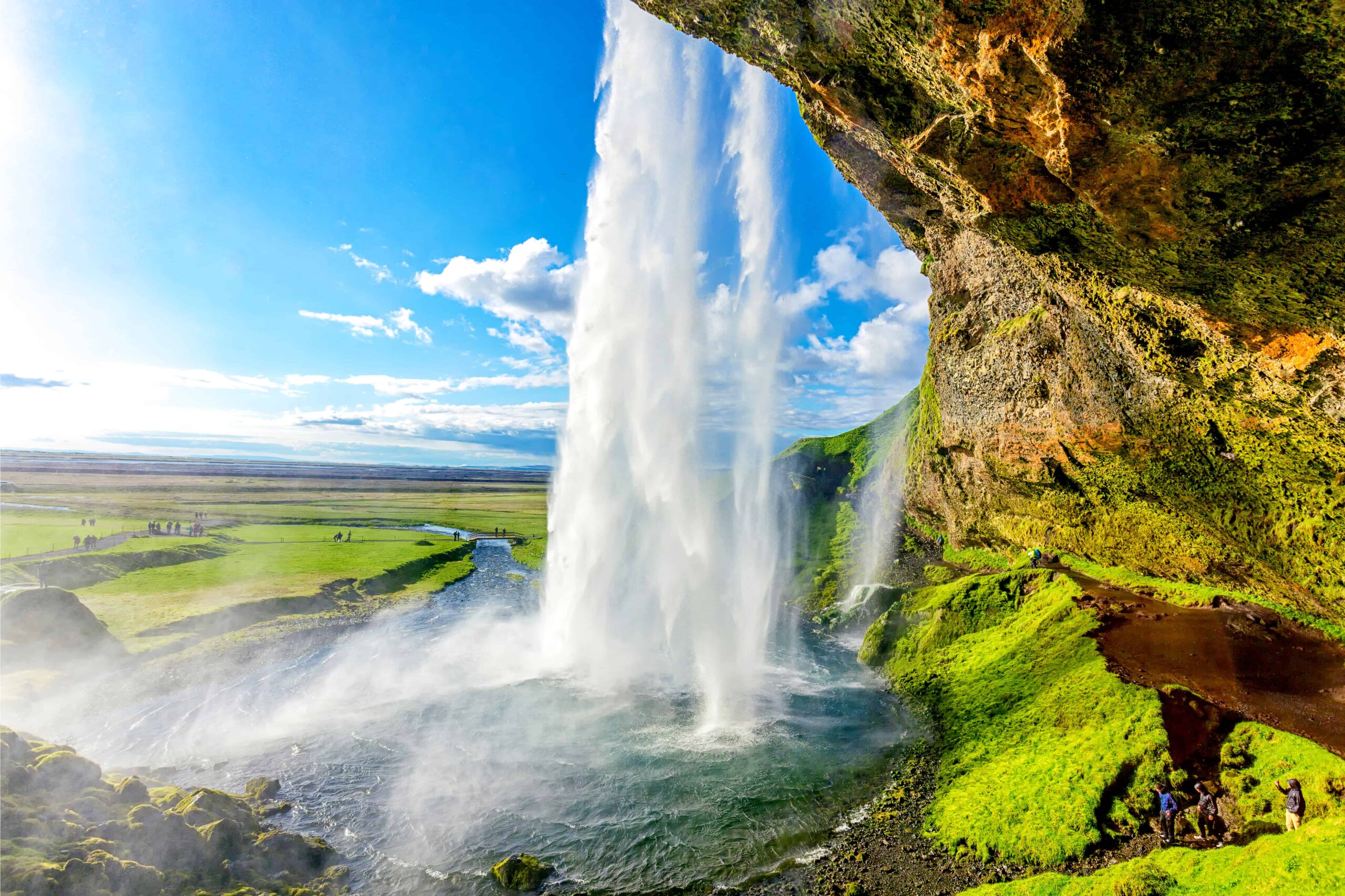 View from behind and beside water that is tumbling over a cliff and then flowing downstream as a river beside green fields. People are on a trail beside the waterfall, looking very small in comparison.