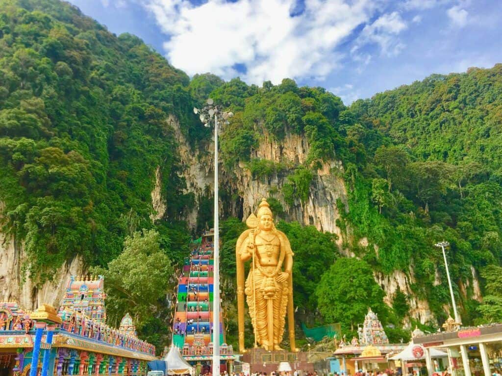 Golden Hindu religious statue outside a cave temple in Malaysia, with limestone cliffs and colorful steps leading to the cave entrance.