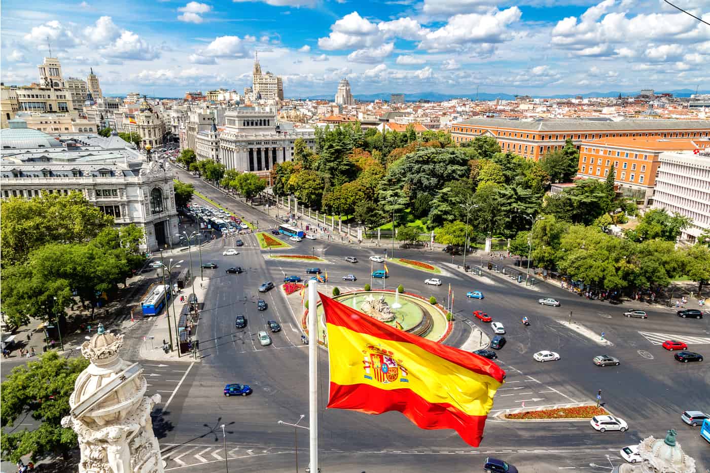 An aerial view of Cibeles Fountain and a large roundabout in the centre of Madrid. In the foreground, a Spanish flag blows in the wind. In the distance, the city stretches out towards the mountains.