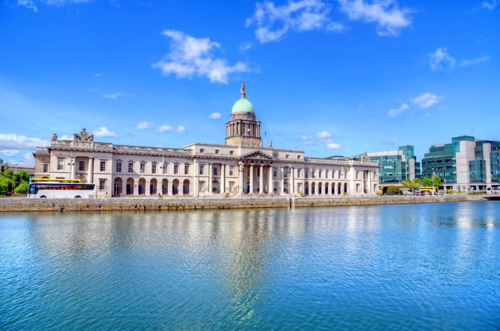 Long, grand stone building with a tower in the centre, stretched along a wide river with buses driving past.