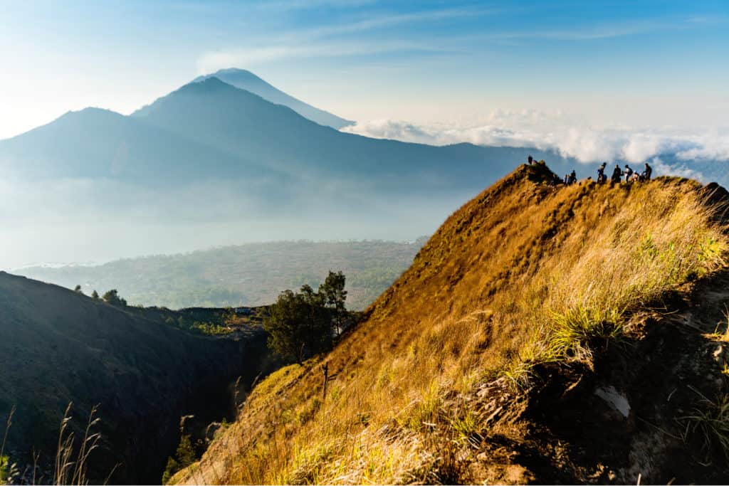 A line of hikers standing  at the top of the narrow ridgeline that marks the summit of Mount Batur in Bali. It's sunrise, and in the distance you can see the silhouettes of volcanos.