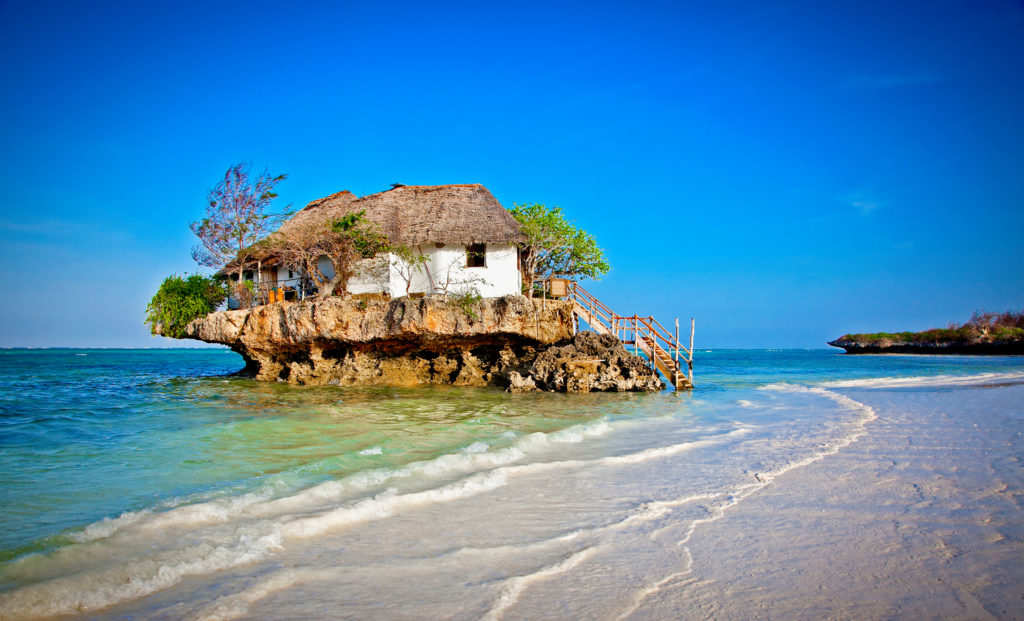 Small rocky island in the ocean shallows with a house built on top of it and wooden stairs from the water.