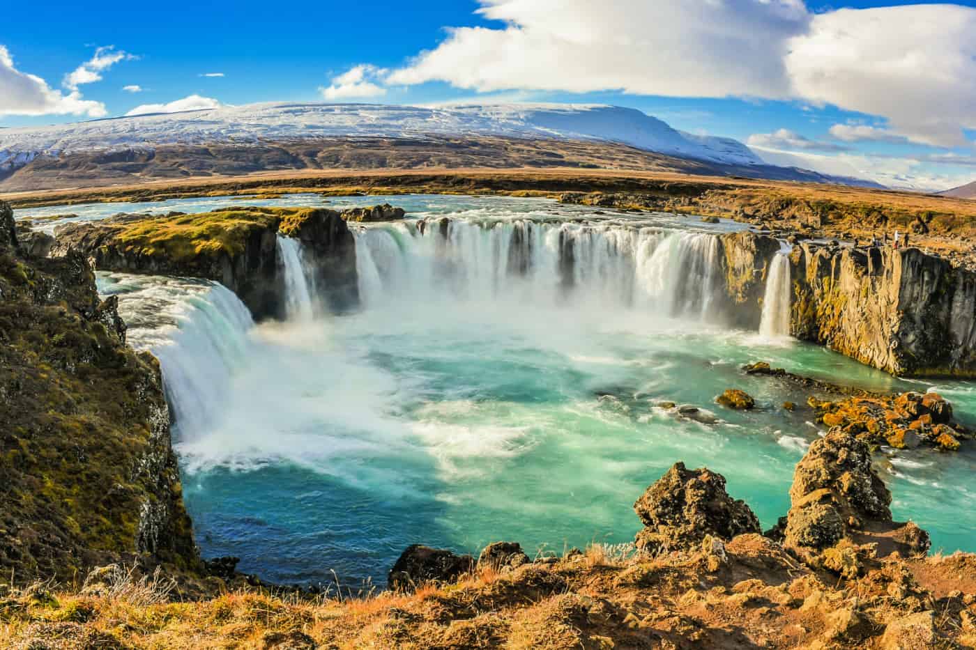 Multiple waterfalls tumbling into a lake below, with snow-covered mountains in the background.