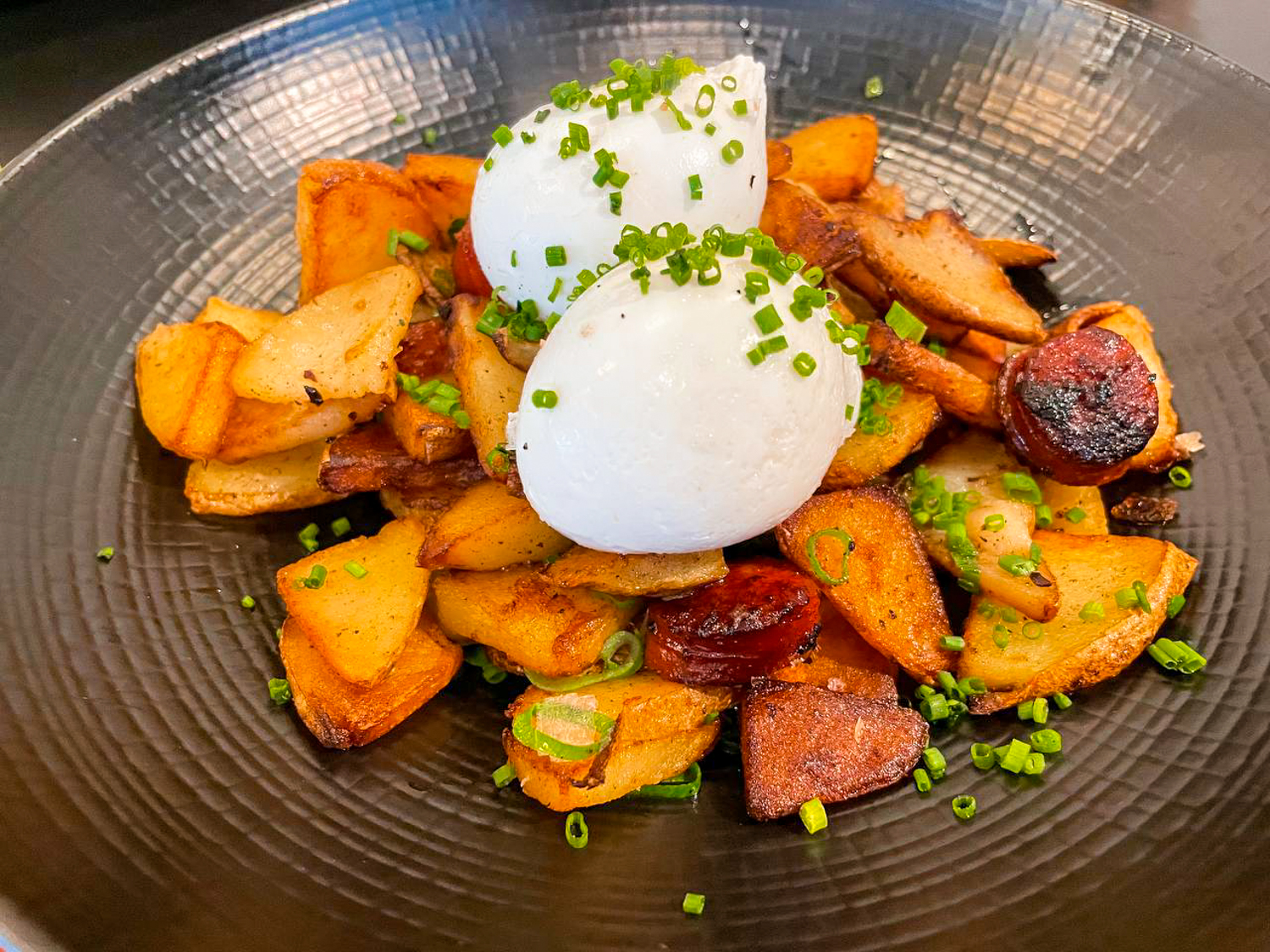A large ceramic plate is filled with a thick layer of roasted potatoes. Charred chorizo, two poached eggs, and a hefty portion of spring onions rest on top of them.