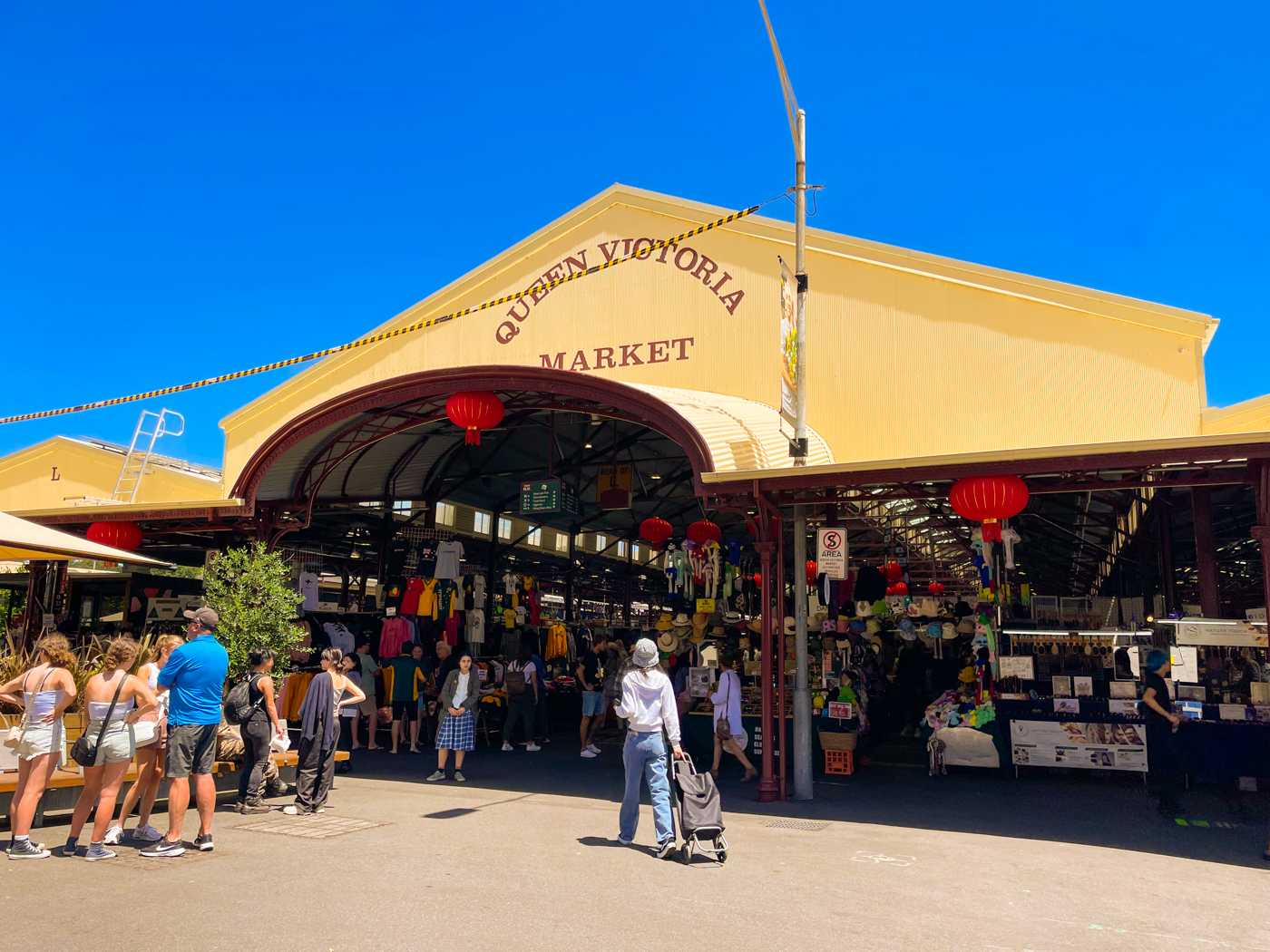The back entrance to the Queen Victoria Market on a cloudless, sunny day. Groups of people are gathering around the open-air building and the interior is lined with clothing and souvenir stalls