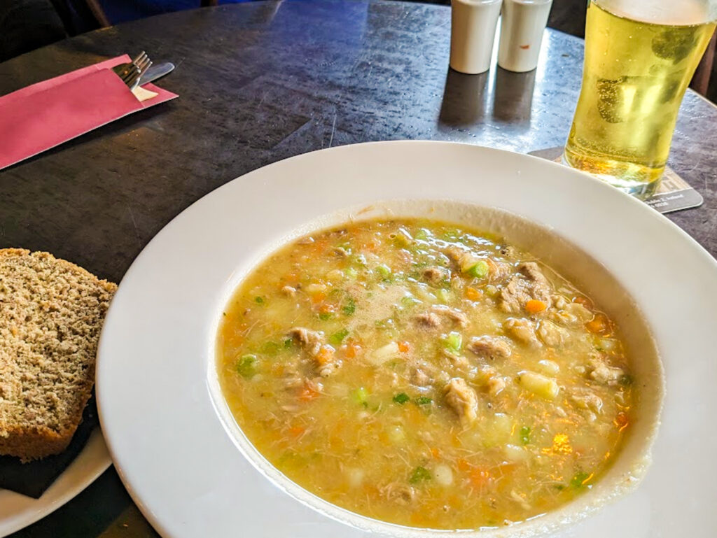 An wooden pub table holds a dish filled with Irish stew. Small chunks of meat and vegetable are visible through the light-coloured broth. To the side is a thick slab of soda bread and in the distance and pint of cider rests.
