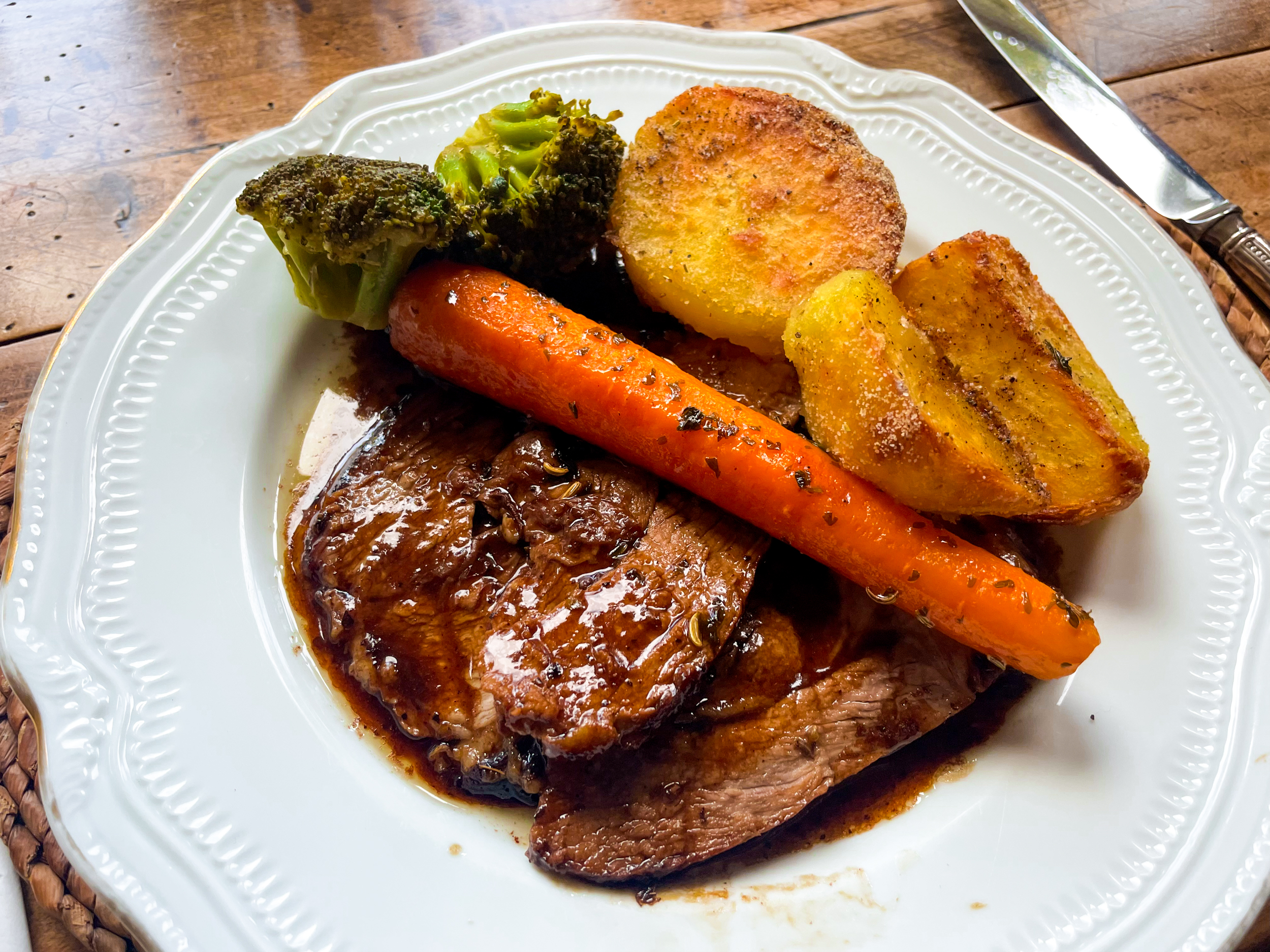A fancy china plate is topped with a high-end version of a roast dinner. Tender slices of beef have been doused in gravy; roasted carrots, broccoli, and potatoes are resting on top of it.