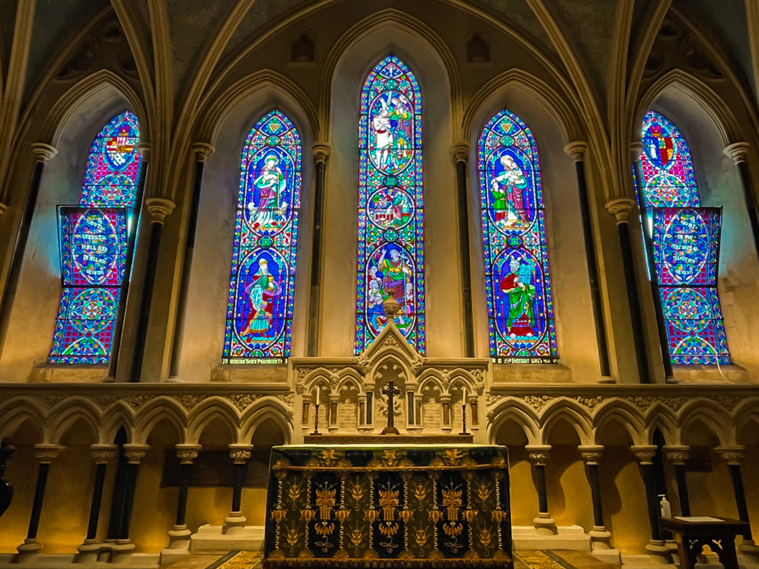 A vast wall of a cathedral that's blanketed in vibrant stained-glass windows, depicting saints and other religious scenes. The lighting is dim yet there's a warmth to the scene.