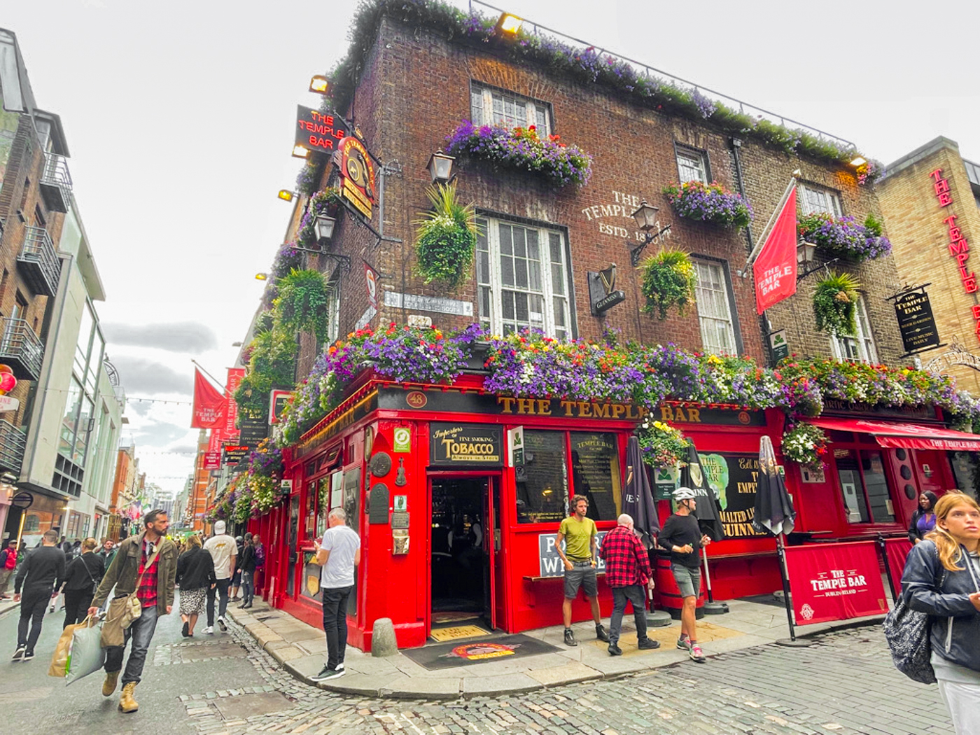The bright red facade of the Temple Bar pub is adorned with hanging baskets, flags, and colourful flowers. The sky is cloudy and grey.